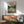 May include: Framed My Neighbor Totoro poster displayed above a wooden sideboard in a mid-century modern living room. The artwork shows children and Totoro character in a meadow with a house in the background, featuring Japanese text. Room includes mint green upholstered chair with wooden arms, small round coffee table, and white ceramic lamp casting natural shadows on white walls.