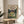 May include: Framed My Neighbor Totoro movie poster displayed on a round wooden side table next to a beige upholstered chair. The poster features the iconic gray Totoro character with children in a green meadow scene, with a cozy house and trees in the background. Soft natural lighting creates gentle shadows across the neutral-toned room setting with a plant visible in the corner.