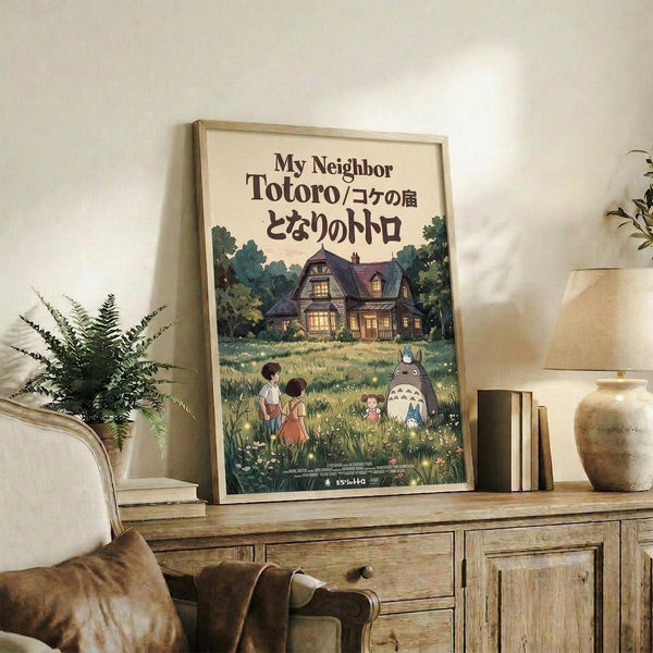 May include: Framed vintage-style movie poster featuring animated characters in a grassy field with a cozy cottage surrounded by trees in the background. The poster displays Japanese text and shows children alongside a large gray forest creature. Set on a rustic wooden sideboard with books, ceramic vase, and fern plant in a warmly lit living room with cream-colored armchair and neutral walls.