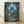 May include: Framed movie poster showing a young girl in red dress and straw hat standing in an enchanted moonlit forest, looking up at the large silhouette of Totoro among glowing fireflies and twisted tree branches. The artwork features deep blues and greens with Japanese text at bottom. The wooden-framed print leans against a white textured wall on light wooden flooring with soft natural lighting creating gentle shadows.