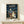 May include: Framed My Neighbor Totoro movie poster displayed on light wooden shelf against white wall. Shows iconic rainy bus stop scene with large gray Totoro holding umbrella, two children with red umbrellas, and smaller blue Totoro. Japanese text above English title. Small wrapped gift box sits in front of frame. Natural lighting creates soft shadows on minimalist setting.