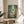 May include: Framed Studio Ghibli My Neighbor Totoro movie poster displaying animated characters in a lush green bamboo forest scene. The poster shows Totoro holding an umbrella with two children and smaller spirits on a woodland path. Wooden frame sits on a round natural wood side table next to a beige upholstered chair, with soft natural lighting and plant shadows creating a cozy living room atmosphere.