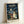 May include: Framed poster of Studio Ghibli's My Neighbor Totoro showing the iconic rainy bus stop scene with Totoro holding an umbrella, two children with red and yellow raincoats, and a smaller blue Totoro character. Japanese text appears at top with English translation below. Dark stormy sky with streetlight creates moody atmosphere. Natural wood frame displayed against white wall corner.