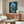May include: Studio Ghibli-inspired poster showing a small child in red dress standing in moonlit forest facing large silhouetted creature among trees. Artwork features deep blue night sky with crescent moon, lush green foliage, and Japanese text. Displayed above mid-century modern walnut credenza in minimalist living room with cream walls, white curved chair, and glass coffee table.