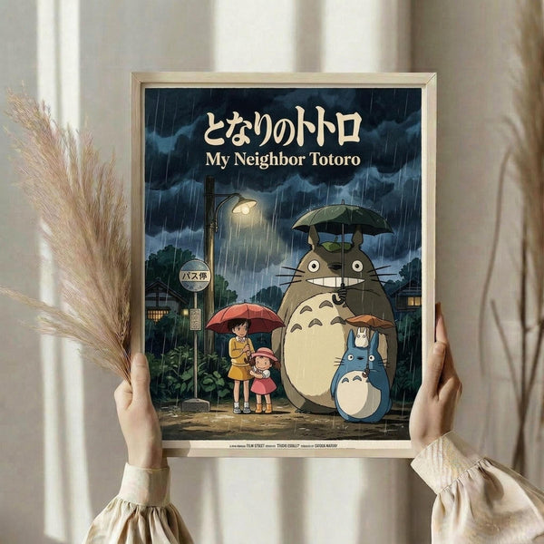 May include: Hands holding a framed poster showing Totoro characters at a rainy bus stop scene with Japanese and English text. The artwork features two children with red umbrellas standing next to large gray forest spirits under an umbrella, with a glowing streetlamp and stormy blue sky. Light wood frame against white curtained window background with dried wheat stems visible on the left side.