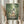 May include: Hands holding a framed Studio Ghibli My Neighbor Totoro movie poster in a light wooden frame. The artwork shows children and Totoro characters walking through a lush green bamboo forest, with the large gray Totoro holding a black umbrella. Dried pampas grass stems are visible on the left side against soft natural lighting and cream curtains in the background.