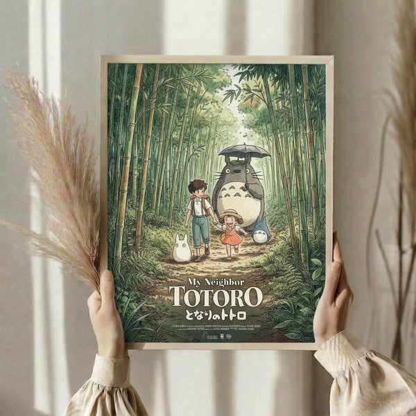 May include: Hands holding a framed Studio Ghibli My Neighbor Totoro movie poster in a light wooden frame. The artwork shows children and Totoro characters walking through a lush green bamboo forest, with the large gray Totoro holding a black umbrella. Dried pampas grass stems are visible on the left side against soft natural lighting and cream curtains in the background.