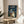 May include: Framed Studio Ghibli My Neighbor Totoro movie poster featuring iconic characters standing in the rain under umbrellas at a bus stop, displayed on a light wood round side table next to a beige upholstered chair with throw pillow, soft natural lighting creating window shadows on neutral wall, small plant visible in corner