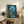 May include: Framed poster featuring animated forest scene with small girl in red dress and hat looking up at large silhouetted creature among trees under crescent moon, displayed on rustic wooden console with books, potted fern, and ceramic vase in cozy living room setting with beige armchair