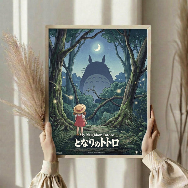 May include: Person holding framed Studio Ghibli-style poster in bright room with pampas grass. Artwork shows small girl in red dress and straw hat looking up at large shadowy Totoro silhouette in moonlit forest. Trees frame the scene with glowing fireflies, crescent moon visible through branches. Poster has light wooden frame, Japanese text at bottom.