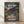 May include: Framed My Neighbor Totoro movie poster featuring two children walking down a twilight village street surrounded by traditional Japanese houses, power lines, and lush green trees. Large glowing eyes peer from the mountainous background. The poster displays both English and Japanese text in white lettering. Warm wooden frame rests against a white wall on light wooden flooring with natural window shadows.