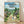 May include: Framed Studio Ghibli My Neighbor Totoro movie poster displayed against white wall on wooden floor. Features iconic characters Totoro, smaller blue Totoro, and white Totoro with two children on dirt path through Japanese village. Background shows traditional houses, mountains, and lush green landscape under blue cloudy sky. Natural wood frame, vintage anime art style with rich colors and detailed countryside setting.