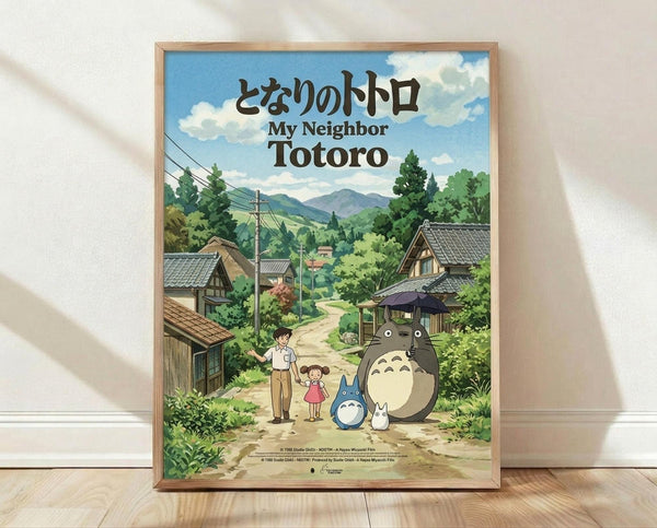 May include: Framed Studio Ghibli My Neighbor Totoro movie poster displayed against white wall on wooden floor. Features iconic characters Totoro, smaller blue Totoro, and white Totoro with two children on dirt path through Japanese village. Background shows traditional houses, mountains, and lush green landscape under blue cloudy sky. Natural wood frame, vintage anime art style with rich colors and detailed countryside setting.