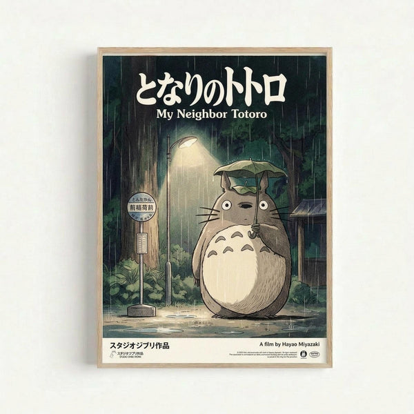 Framed poster featuring Totoro, the beloved gray forest spirit character, standing at a rainy bus stop while holding a small umbrella. The scene shows heavy rain falling, a wooden bus stop sign, lush green vegetation, and atmospheric lighting. Japanese text appears at the top with "My Neighbor Totoro" in English below, and "A film by Hayao Miyazaki" at the bottom. The poster has a nostalgic, hand-drawn animation art style with muted earth tones and is displayed in a light wooden frame ag