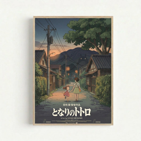 Framed poster showing an illustrated scene of two children walking hand-in-hand down a rural Japanese street at dusk. The artwork features traditional wooden houses with tiled roofs, power lines, lush green trees, and mountains in the background under a sunset sky. Japanese text appears at the bottom along with "A film by HAYAO MIYAZAKI" in English. The poster has a light wooden frame and is mounted on a white wall.