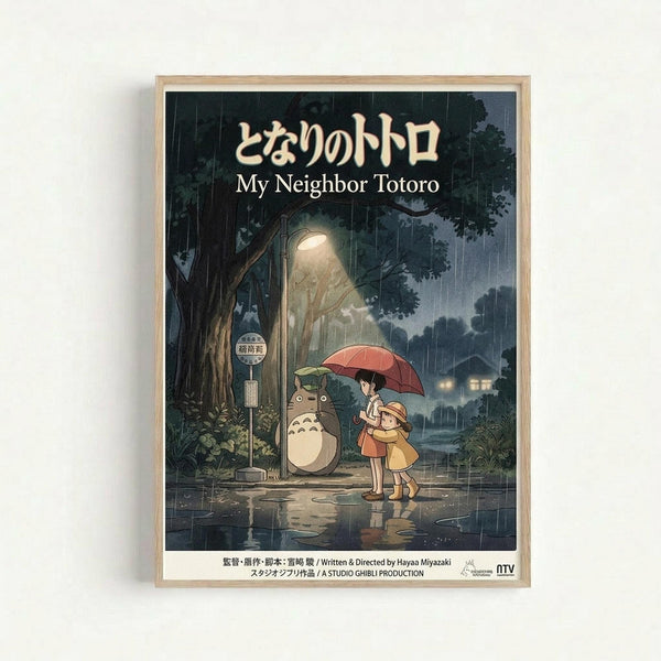 Framed poster featuring animated characters from Studio Ghibli's My Neighbor Totoro standing at a rainy bus stop. Two children with a red umbrella wait beside the large gray forest spirit Totoro under a glowing street lamp. The scene shows a nighttime forest setting with heavy rain, puddles reflecting light, and a bus stop sign. Japanese and English text appears at the top, with production credits at the bottom. The poster has a warm wooden frame against a white wall.
