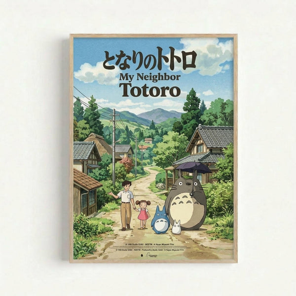 Framed poster featuring the iconic Studio Ghibli characters Totoro in a peaceful Japanese countryside village scene. The artwork shows a large gray Totoro with smaller blue Totoro and white spirit, alongside two children on a dirt path surrounded by traditional wooden houses, lush green trees, rolling hills, and power lines under a cloudy blue sky. The poster has Japanese text at the top with "My Neighbor Totoro" in English below, displayed in a light wooden frame against a white background.