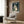 May include: Studio Ghibli My Neighbor Totoro movie poster mounted on wood-paneled wall above beige upholstered armchair with geometric patterned pillow. Poster shows iconic Totoro character standing at rainy bus stop with umbrella in dark green tones. Modern living room setting with brass floor lamp, side table with open book, and natural lighting from window with curtains.