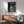 May include: Framed My Neighbor Totoro movie poster mounted on white wall above wooden sideboard with white table lamp. Poster shows Totoro and two children with red umbrella at rainy bus stop in forest. Mid-century modern living room with sage green armchair, round coffee table, and natural sunlight streaming across scene.