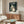 May include: Framed My Neighbor Totoro movie poster featuring the iconic gray forest spirit holding an umbrella in the rain, displayed above a mid-century modern wooden credenza with sliding doors. Scene shows white upholstered chair, oval glass coffee table, and minimalist floor lamp in neutral-toned living room with natural lighting.