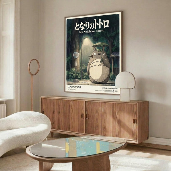 May include: Framed My Neighbor Totoro movie poster featuring the iconic gray forest spirit holding an umbrella in the rain, displayed above a mid-century modern wooden credenza with sliding doors. Scene shows white upholstered chair, oval glass coffee table, and minimalist floor lamp in neutral-toned living room with natural lighting.