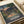 May include: Studio Ghibli movie poster print displayed on white fabric surface next to rolled paper tube. Vintage-style artwork shows two children walking on rural path between traditional Japanese houses at dusk, with mountains and power lines in background. Japanese text at bottom reads "となりのトトロ" with "A film by HAYAO MIYAZAKI" credit. Warm sunset colors blend orange and purple skies with detailed animation-style illustration in earth tones.