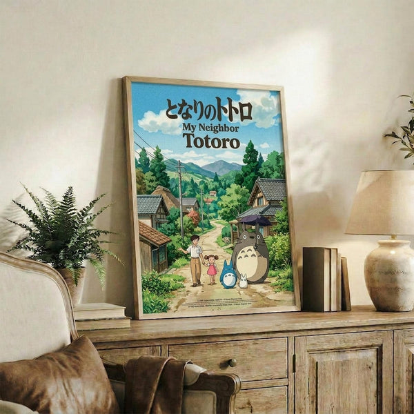 May include: Framed Studio Ghibli My Neighbor Totoro movie poster displayed on rustic wooden console table in cozy living room setting. Poster shows Japanese village scene with iconic gray Totoro character, young children, traditional houses, and lush green landscape under blue sky. Natural wood frame complements surrounding decor including fern plant, books, ceramic vase, and neutral-toned furniture against white wall.