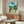 May include: My Neighbor Totoro movie poster displayed above a wooden sideboard in a modern living room setting. The colorful animated poster shows Totoro and characters in a rural Japanese village scene with mountains and green trees. White upholstered chair and glass-top oval coffee table visible in foreground. Warm wood tones throughout the minimalist interior space with cream walls and natural lighting.