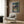 May include: Framed Studio Ghibli My Neighbor Totoro movie poster displayed on wood-paneled wall above modern cream armchair with geometric pillow. Artwork shows two children walking through Japanese village at dusk with traditional houses, trees, and power lines. Warm interior lighting with gold floor lamp and dark round side table with open book.