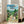 May include: Framed Studio Ghibli poster showing animated characters Totoro and smaller blue spirit with two children on a dirt path through a lush green Japanese village. Rolling mountains and traditional houses with tiled roofs fill the background under a bright blue cloudy sky. The poster leans against a white wall next to a beige upholstered chair in a sunlit room with large windows and light wooden floors.