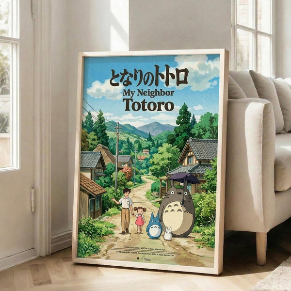 May include: Framed Studio Ghibli poster showing animated characters Totoro and smaller blue spirit with two children on a dirt path through a lush green Japanese village. Rolling mountains and traditional houses with tiled roofs fill the background under a bright blue cloudy sky. The poster leans against a white wall next to a beige upholstered chair in a sunlit room with large windows and light wooden floors.