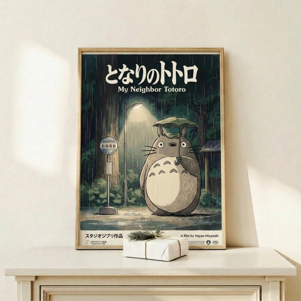 May include: Framed My Neighbor Totoro movie poster in natural wood frame, displayed on light wooden shelf against cream wall. Features iconic Totoro character holding umbrella in rain at bus stop, with Japanese text and Studio Ghibli styling in muted greens and grays. Small wrapped gift box with pine sprig sits beside frame.