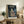 May include: Framed Studio Ghibli movie poster featuring Totoro and two children with red umbrella standing at rainy bus stop under large tree. Poster displays in light wood frame on rustic wooden console table beside cream armchair, fern plant, and ceramic vases in cozy living room setting with neutral walls.