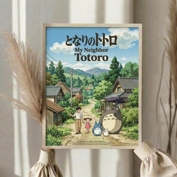 May include: Framed poster displaying animated scene with large gray forest creature, two children, and smaller blue creature on dirt path through Japanese village. Green mountains and traditional houses with tiled roofs in background under blue cloudy sky. Japanese and English text at top. Light wooden frame held by hands wearing cream ruffled sleeves against soft beige curtained background with dried wheat stalks.
