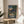 May include: Framed Studio Ghibli movie poster featuring two children walking down a rural Japanese village street at dusk, with traditional houses, power lines, and mountains in the background. The artwork shows warm sunset colors and Japanese text at the bottom. The wooden-framed print sits on a round light wood side table next to a beige upholstered chair, with natural window light casting shadows and a plant partially visible.