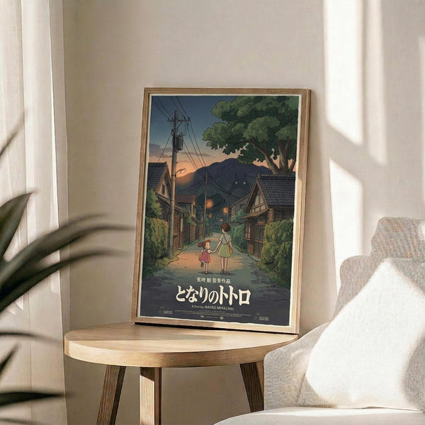 May include: Framed Studio Ghibli movie poster featuring two children walking down a rural Japanese village street at dusk, with traditional houses, power lines, and mountains in the background. The artwork shows warm sunset colors and Japanese text at the bottom. The wooden-framed print sits on a round light wood side table next to a beige upholstered chair, with natural window light casting shadows and a plant partially visible.