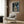 May include: Framed My Neighbor Totoro movie poster mounted on wood-paneled wall above modern beige armchair with geometric pillow. Poster shows Totoro and children with umbrella at rainy bus stop scene in muted blues and greens. Room features gold floor lamp, black side table with open book, and neutral contemporary decor with warm wood tones throughout.