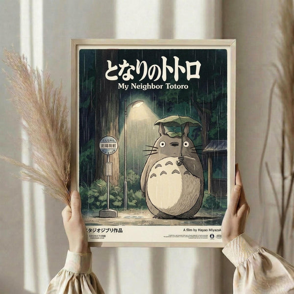 May include: Hands holding a framed vintage-style movie poster featuring Totoro, the beloved animated character, standing at a rainy bus stop while holding an umbrella. The poster displays Japanese text at the top with English translation below, set against a moody forest scene with falling rain. A person in cream-colored clothing with ruffled sleeves holds the light wood-framed artwork, with dried wheat stalks visible to the left side of the soft, neutral-toned room setting.