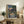 May include: Framed Studio Ghibli movie poster showing two children walking down a rural Japanese street at dusk, with traditional houses, power lines, and mountains in background. Poster displays in light wood frame on rustic wooden sideboard alongside fern plant, books, and ceramic vase in cozy living room setting with beige armchair.