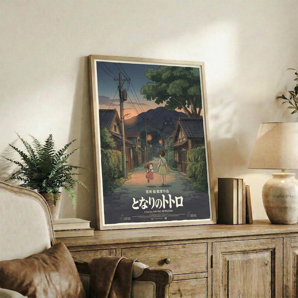 May include: Framed Studio Ghibli movie poster showing two children walking down a rural Japanese street at dusk, with traditional houses, power lines, and mountains in background. Poster displays in light wood frame on rustic wooden sideboard alongside fern plant, books, and ceramic vase in cozy living room setting with beige armchair.