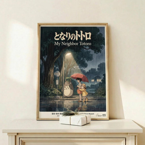 May include: Framed poster of Studio Ghibli's My Neighbor Totoro showing two children with red umbrella standing at rainy bus stop beside large gray Totoro character. Dark moody forest scene with glowing lights in background. Natural wood frame displayed on light wooden shelf with small wrapped gift box. Warm beige wall behind.
