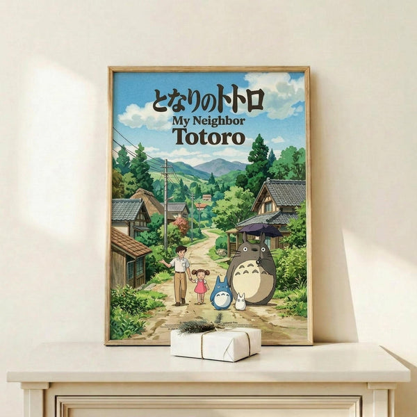 May include: Framed poster displaying animated rural Japanese village scene with rolling green hills, traditional houses, and iconic gray forest creature alongside smaller blue companion and two children on dirt path. Wooden frame sits on light wooden shelf next to wrapped white gift box with small plant sprig. Soft natural lighting against cream wall.
