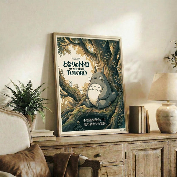 May include: Framed My Neighbor Totoro poster displayed on rustic wooden console table in living room setting. The illustrated poster shows the beloved grey forest spirit sitting peacefully in a lush tree with Japanese text. Natural wood frame complements the earthy artwork. Scene includes fern plant, ceramic vase, books, and beige armchair, creating a cozy home interior with warm natural lighting and neutral wall colors.