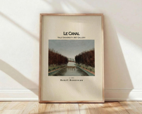 May include: Framed art print leaning against white wall on light wood floor. Cream-colored poster features a painting of a canal lined with bare winter trees under an overcast sky. Text reads "Le Canal" and "Yale University Art Gallery" above the artwork, with "Henri Rousseau" below. Natural wood frame with soft window light casting shadows across the scene.