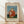 May include: Framed art print displaying Henri Rousseau's 1906 painting of a blonde child in a red polka-dotted dress holding a small doll, set against a blue sky background. The wooden frame sits on light wood flooring against a white wall with natural sunlight casting diagonal shadows across the scene.