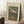 May include: A framed vintage botanical poster leaning against white walls on a textured cream rug. The artwork shows colorful flowers including pink peonies, white daisies, and red tulips in a ribbed glass vase against a teal green background with an orange banner below. A white coffee cup sits on a wooden block nearby, with soft natural lighting creating shadows on the wall.