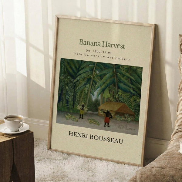 May include: Framed art poster leaning against white wall showing tropical jungle scene with banana trees, small figures carrying bundles, and thatched huts. Cream-colored mat with gallery text and artist name Henri Rousseau below the artwork. Natural wood frame rests on textured white rug beside wooden side table with coffee cup and beige throw blanket in bright, minimalist room setting.