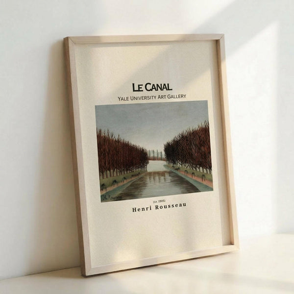 May include: Framed art print of Henri Rousseau's "Le Canal" painting showing a waterway lined with bare trees under a gray sky. The vintage-style poster features cream background with black text identifying the artwork and Yale University Art Gallery. Light wood frame displayed against white wall, leaning on surface with soft natural lighting casting gentle shadows.