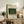 May include: Framed vintage art poster featuring Henri Rousseau's tropical jungle scene with banana trees and small figures, displayed on rustic wooden console alongside fern plant, ceramic vase, and books in neutral-toned living room setting