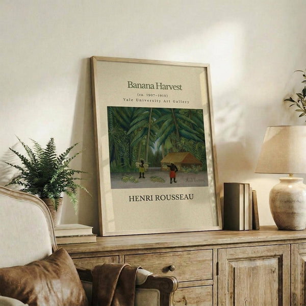 May include: Framed vintage art poster featuring Henri Rousseau's tropical jungle scene with banana trees and small figures, displayed on rustic wooden console alongside fern plant, ceramic vase, and books in neutral-toned living room setting